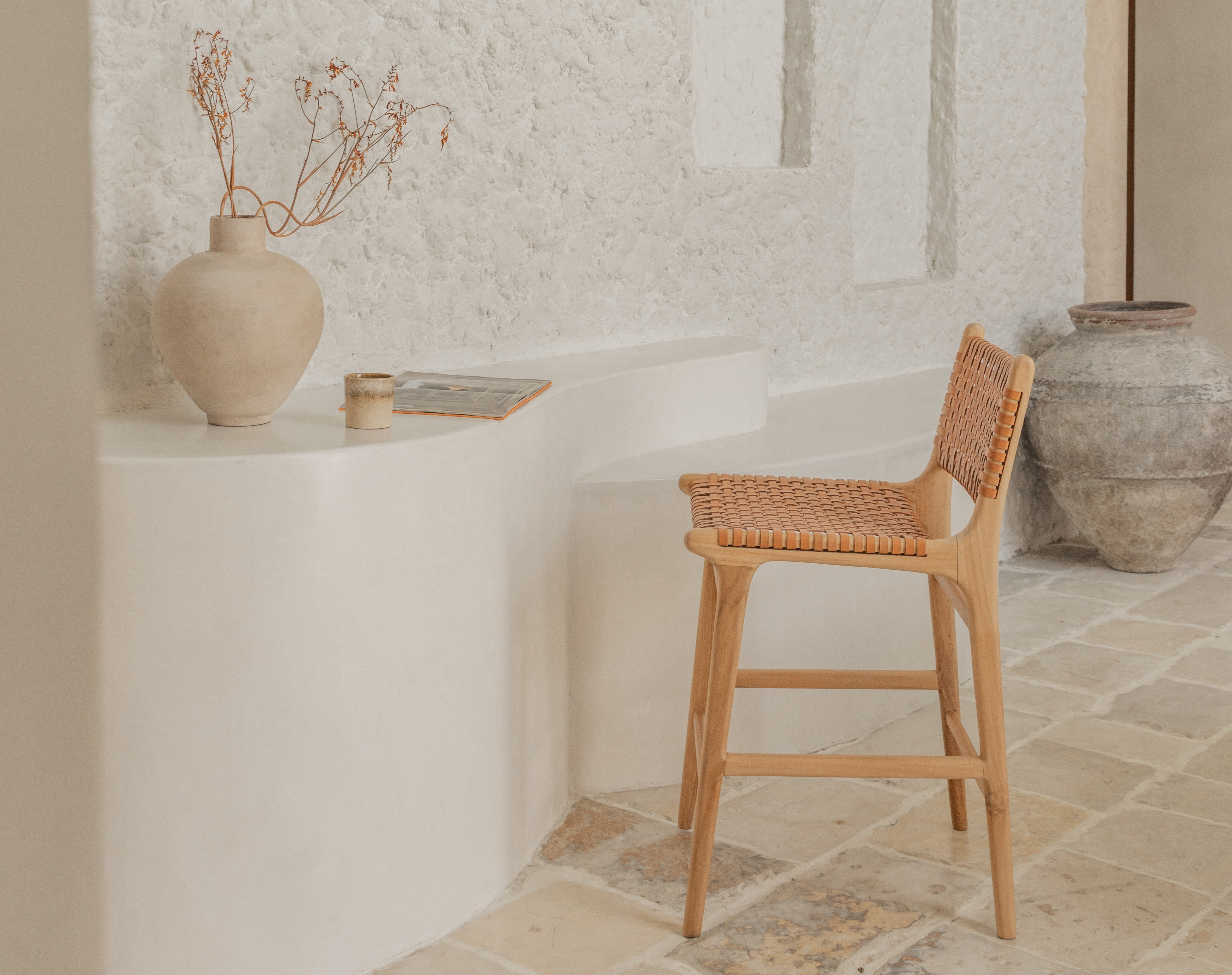 Troffle handcrafted teak and woven leather bar stool in a warm minimalist interior with stone floor and textured white walls